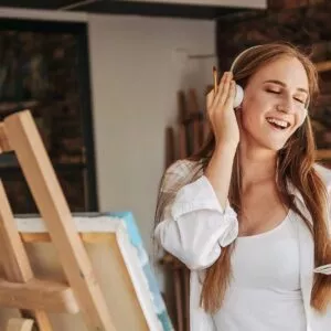 Smiling young lady listening to music and painting picture, enjoying art therapy