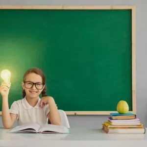 A schoolgirl holds in her hand a lamp with light sitting at a desk in school.